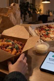 Rigatoni in der Takeaway-Box, Pizza im Karton und ein Smartphone mit Bestellbestätigung auf einem Esstisch am Abend