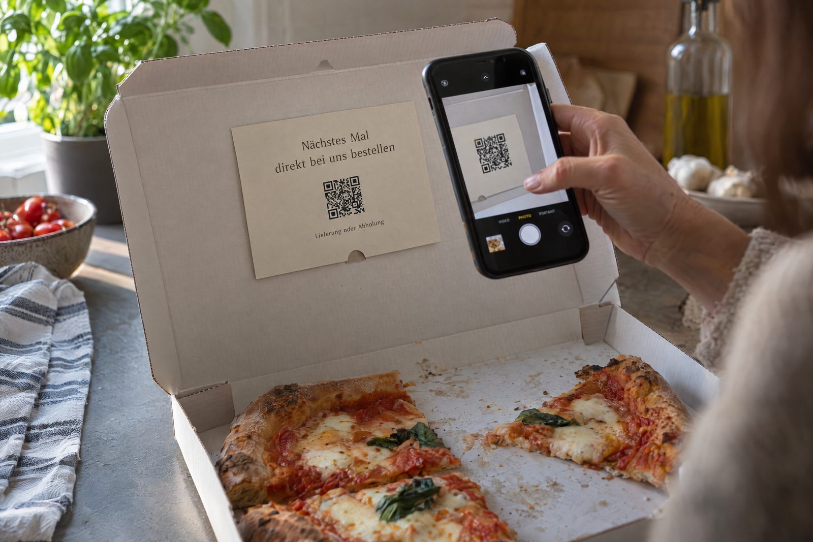 Karte mit QR-Code und dem Text Nächstes Mal direkt bei uns bestellen im Deckel eines Pizzakartons, eine Hand scannt sie mit dem Smartphone